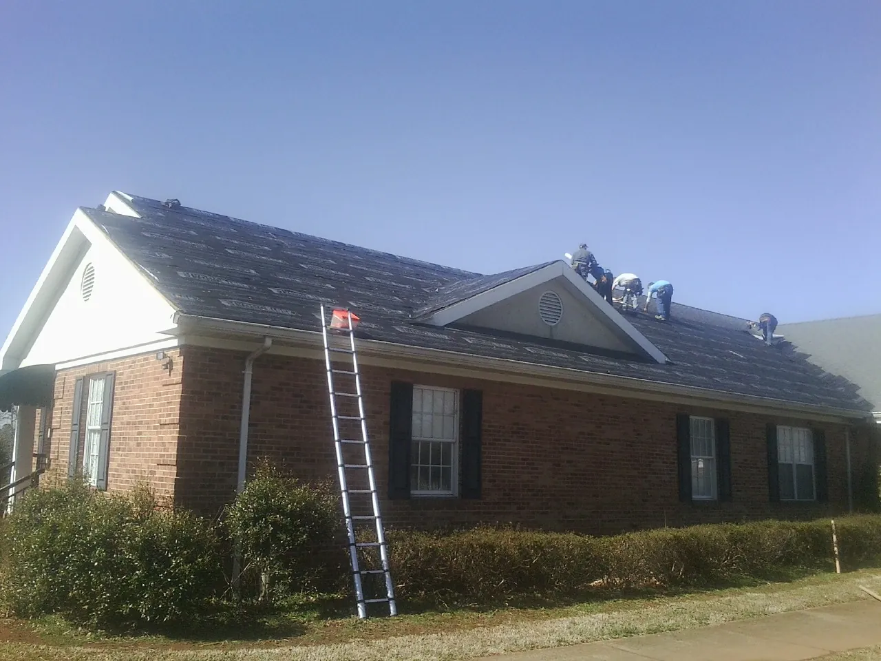 Crew tearing shingles off a brick ranch roof with a ladder set — Emergency Roof Repair work by our crew in Angleton